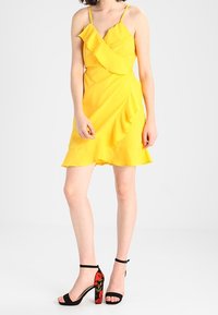 Gelbes knielanges Kleid mit Rüschendesign und dünnen Trägern. Kombiniert mit schwarzen High-Top-Sandalen mit lebendigen floralen Details.