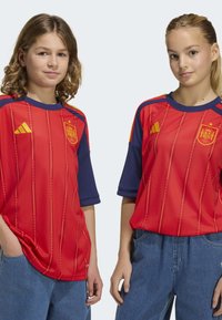 Röda fotbollströjor med gula detaljer och blå ärmar. Designelement inkluderar vertikala ränder, lagets emblem på bröstet och Adidas-logotyp.