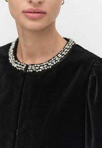 Czarny aksamitny strój z okrągłym dekoltem, ozdobiony metaliczną i kryształową koronką z koralików. Poszerzane rękawy dla efektu tekstury.