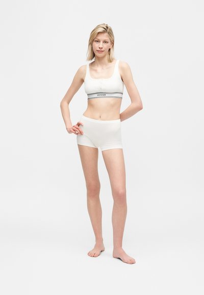Γυναίκα που φοράει λευκό αθλητικό σουτιέν με το λογότυπο Versace και αντίστοιχα λευκά σορτς με ψηλή μέση, όρθια και ξυπόλυτη σε λευκό φόντο.