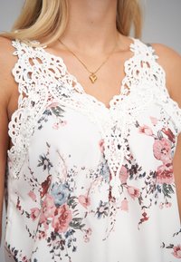 Bloes met bloemenpatroon in witte stof, met roze en blauwe bloemen. Kant rondom de halslijn geeft structuur. Accessoire: gouden hartvormige ketting.