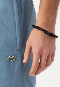 Blauwe lichtgewicht stoffen shorts met een groen geborduurd krokodillenlogo aan de zijkant; een zwarte schakelarmband om de pols.