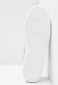 Suola di sneaker bianca con sezioni di presa testurizzate e marchi del brand, posizionata verticalmente contro un muro bianco nell'angolo.