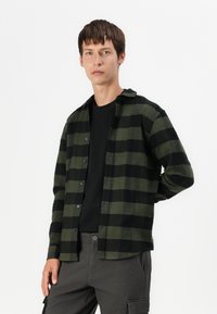 Zwart en groen geruit flanellen shirt met lange mouwen, knoopsluiting en een rechte snit, boven een zwart T-shirt.