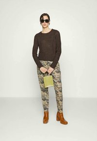 Mulher vestindo um suéter castanho escuro, calças camufladas, botas de salto castanhas, óculos de sol pretos grandes, segurando uma pequena bolsa verde oliva.