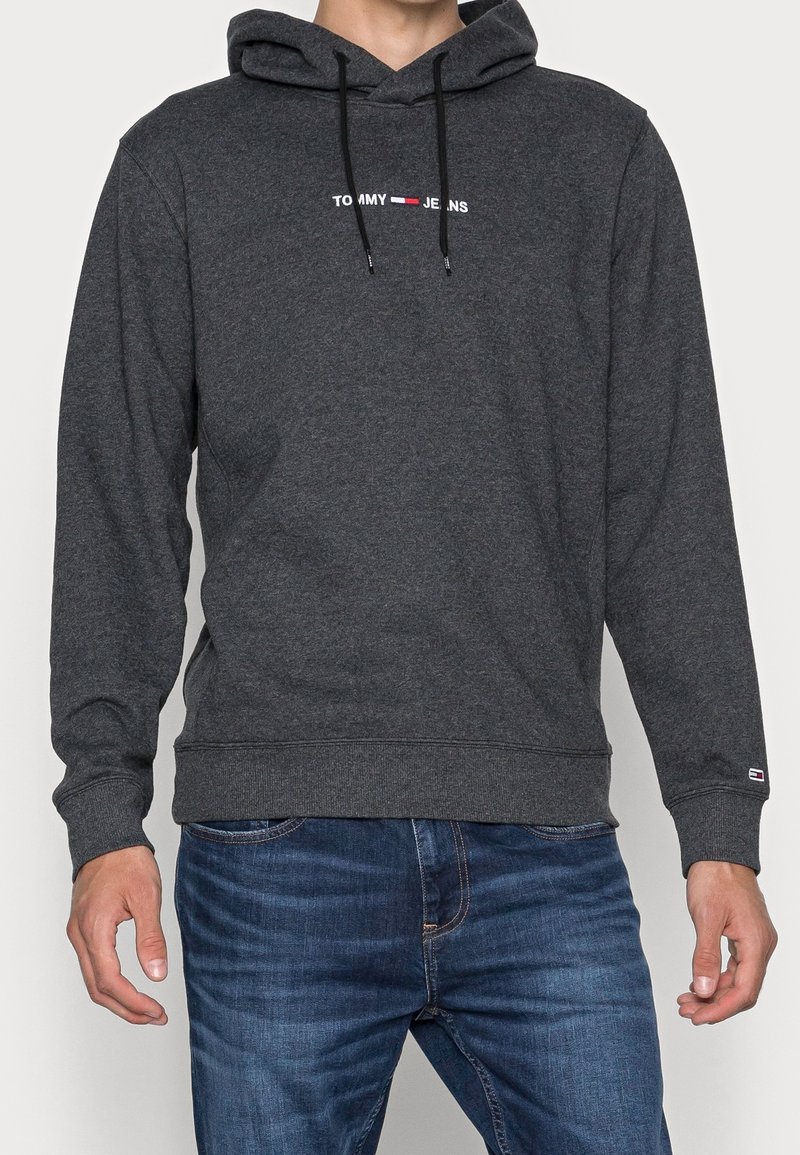Homme portant un sweat à capuche Tommy Jeans gris foncé et un jean bleu, debout avec les bras détendus le long du corps devant un fond uni.