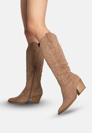 Gambe con stivali da cowboy in camoscio color tan fino al polpaccio, con motivi ricamati, cerniere laterali e tacchi in legno massiccio, su uno sfondo chiaro e semplice.