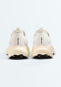 Paire de chaussures de sport beiges avec des semelles épaisses et des bandes réfléchissantes verticales sur le talon, vues de dos sur un fond uni.