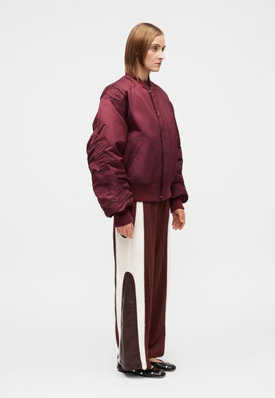 Veste bomber matelassée bordeaux avec poignets en côte, associée à un pantalon large avec panneaux blancs et marron, et des chaussures plates noires.
