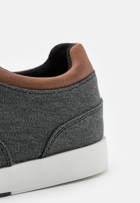 Grå lerrets sneakers med en brun lærhælt accent, teksturert stoff og hvit gummisåle. Nærbilde viser detaljerte sømmer.