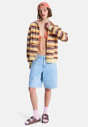 Pruhovaná žlto-hnedá mikina s kapucňou, oranžový bikini top, svetlomodré denimové šortky, ružové ponožky, hnedé sandále a ružová čiapka.