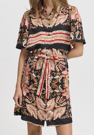 Vrouw die een korte mouwen, met knopen gesloten jurk draagt met een rood, zwart en gouden ingewikkeld paisley- en bloemenpatroon, gestrikt in de taille met een bijpassende riem.