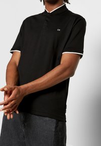 Polo noir avec col, doté d'une patte de boutonnage à trois boutons et de finitions blanches sur les manches. Confectionné dans un tissu lisse et léger.