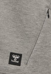 Tissu de sweat-shirt gris avec une finition texturée et une fermeture éclair sur le côté. Patch de logo de marque rectangulaire noir avec des détails blancs dans le coin inférieur.