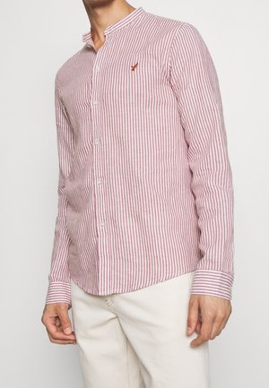 Muž nosící dlouhý rukáv, červenobíle vertikálně pruhovanou košili na knoflíky s malým vyšitým logem na hrudi, v kombinaci se světlými kalhotami.