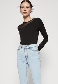 Mlada žena nosi crnu majicu dugih rukava s prozirnim izrezom i svijetlo plave traperice visokog struka, stoji s jednom rukom u džepu.