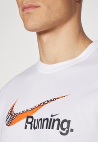 Camiseta de algodón blanca con el texto "Running." en negro y un logo de swoosh de Nike multicolor que presenta acentos en naranja y patrones.