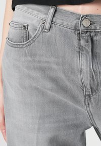 Grijze denim jeans met een klassiek vijf-pocket ontwerp, metalen hardware accenten en zichtbare stikdetails. De stof heeft een subtiele textuur.