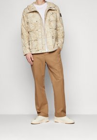 Ljust beige jacka med ett kartliknande mönster, flera fickor och huva. Bärs över en vit t-shirt och är ihopparad med beige byxor och krämfärgade sneakers.