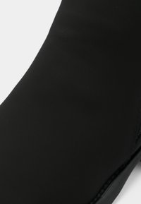 Tuvplāns ar gludu melnu apavu augšdaļu, redzams minimāls šuvums pie zolītes malas uz balta fona.