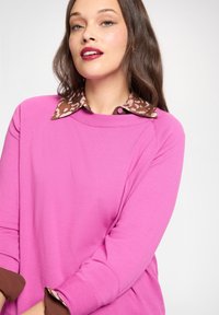Różowy sweter typu pulower z okrągłym dekoltem, kontrastującym brązowym kołnierzem w panterkę i brązowymi mankietami. Gładka tkanina o luźnym kroju.