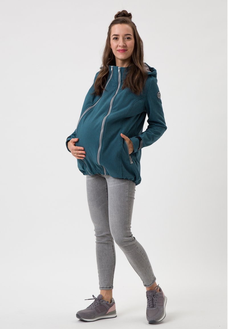 Türkise Umstandjacke mit Kapuze, doppeltem Vorderreißverschluss und Seitentaschen, kombiniert mit hellgrauen Jeans und grauen Sportschuhen.