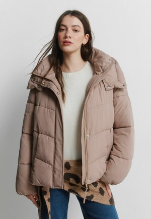 Jonge vrouw met een beige gewatteerde jas over een witte trui en een sjaal met luipaardprint, poserend tegen een effen lichte achtergrond.