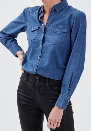 Kobieta w niebieskiej, zapinanej na guziki koszuli z długim rękawem i kieszeniami na piersi oraz czarnych spodniach z wysokim stanem, z ręką na biodrze.