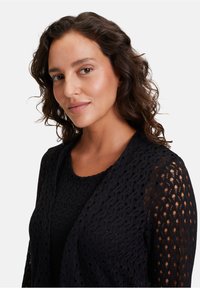 Schwarze offene Strickjacke mit strukturiertem Muster und langen Ärmeln, die über einem einfarbigen schwarzen Top getragen wird, aus leichtem, atmungsaktivem Stoff.