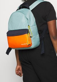 Persona che indossa un completo nero con uno zaino a blocchi di colore in azzurro, blu navy e arancione, dotato di estrattori gialli sulla parte posteriore.
