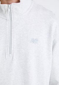 Felpa grigia chiara con zip realizzata in tessuto morbido, dotata di collo alto e con un logo "N" blu chiaro ricamato sul petto.