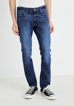 Jean bleu foncé en denim, avec une coupe ajustée, un design à cinq poches et des coutures jaunes contrastantes, portés avec des baskets noires.