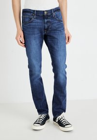 Mørkeblå jeans laget av denim, med slim-fit, fem-lommers design og kontrasterende gul søm, brukt med svarte sneakers.