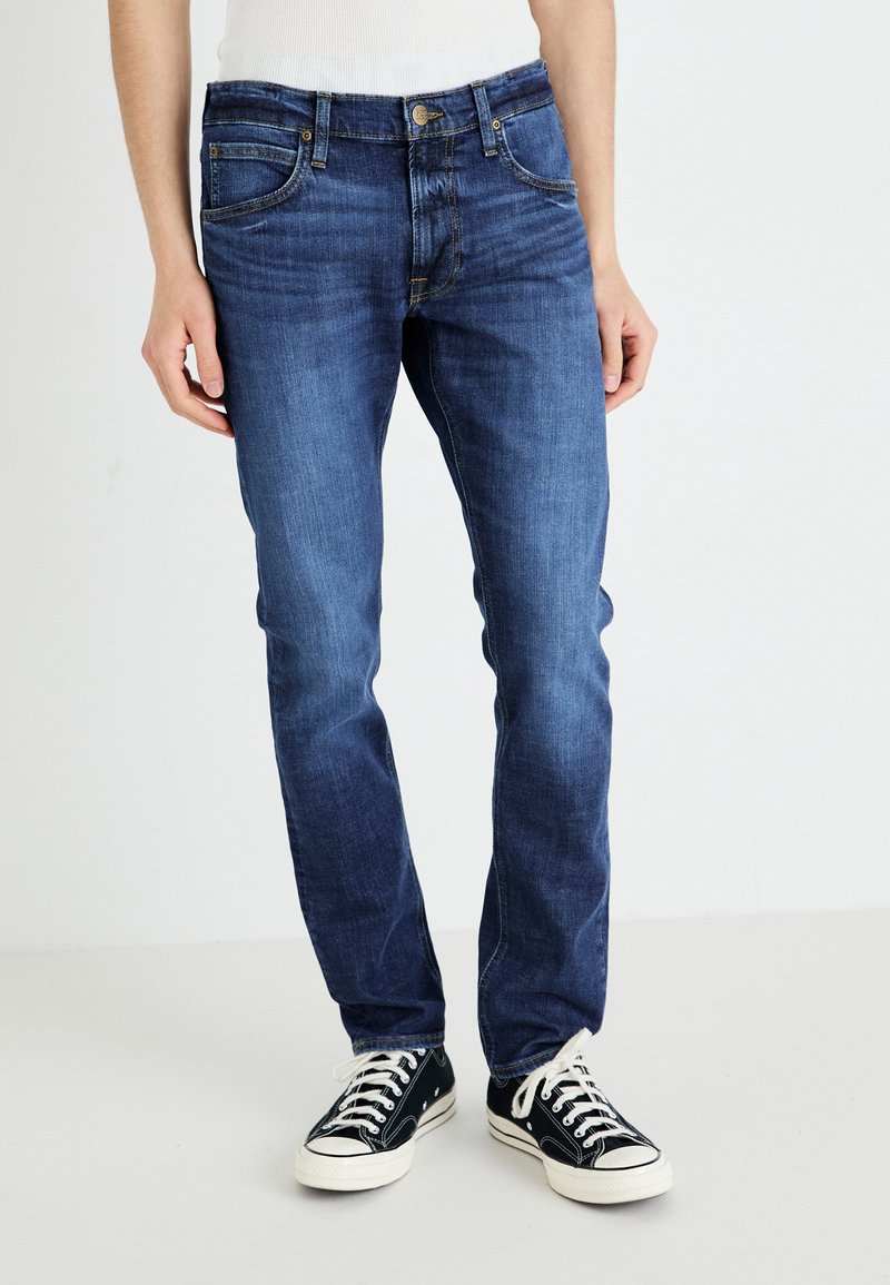 Mørkeblå jeans laget av denim, med slim-fit, fem-lommers design og kontrasterende gul søm, brukt med svarte sneakers.