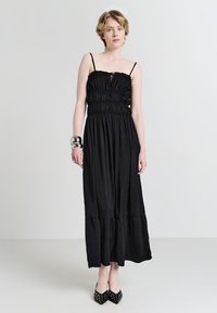 Schwarzes Maxikleid mit elastischem Oberteil, dünnen Trägern und einem gestuften Saum. Kombiniert mit spitzen schwarzen Schuhen mit Nietenakzenten.