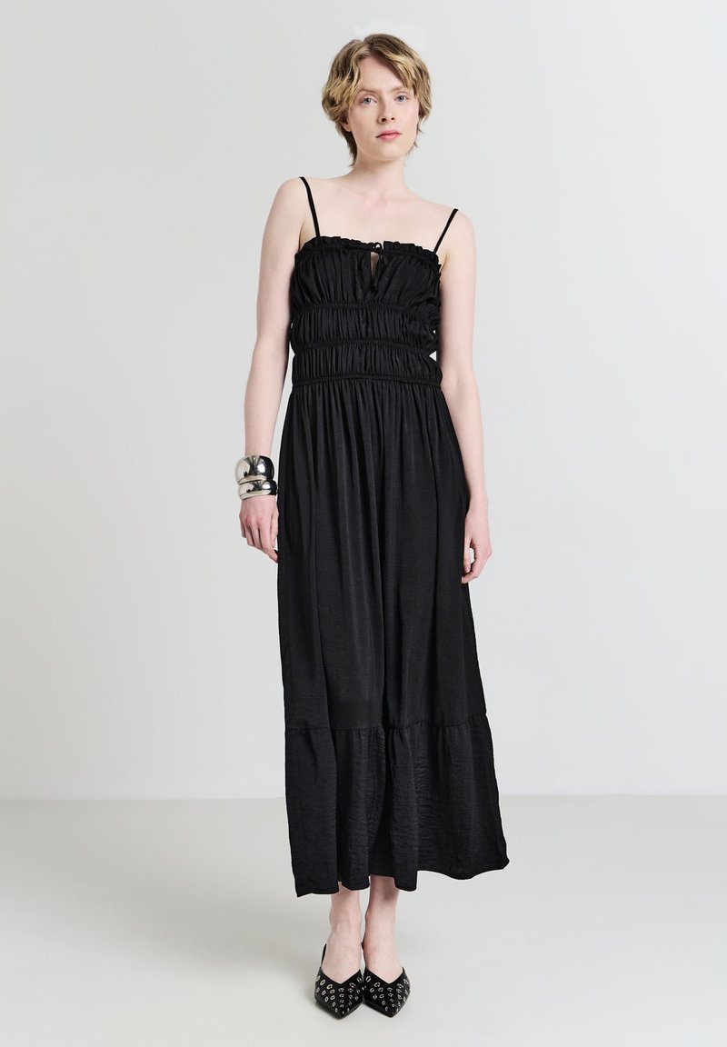 Schwarzes Maxikleid mit elastischem Oberteil, dünnen Trägern und einem gestuften Saum. Kombiniert mit spitzen schwarzen Schuhen mit Nietenakzenten.