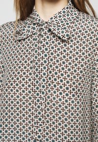 Femme portant un chemisier boutonné avec un grand col et un imprimé floral géométrique dans des tons sarcelle, marron et beige.