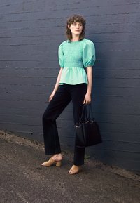 Top em verde menta com mangas bufantes e corpete franzido, combinado com calças pretas de boca de sino e mules bege. A modelo segura uma bolsa preta com alças.