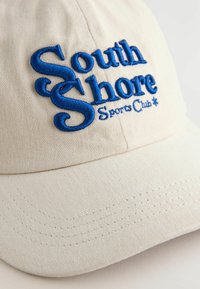Casquette de baseball couleur crème en tissu texturé, avec un texte bleu brodé qui dit "South Shore Sports Club."