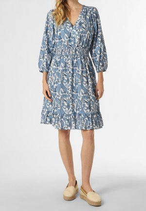 Kvinde iført en blå og hvid knælang kjole med paisleymønster, lange ærmer, indsnøret talje, flæsekant og beige platform slip-on sko.