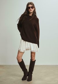 Hnedý oversized sveter, biele šaty s čipkovým lemom a hnedé semišové čižmy po kolená. Modelka má na sebe veľké slnečné okuliare; hladké a štruktúrované materiály.