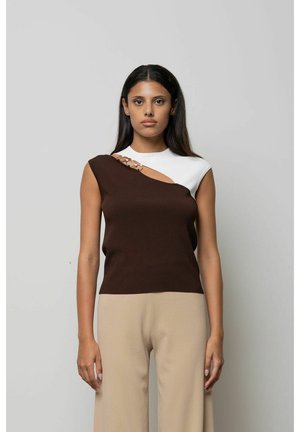 Donna con capelli lunghi e scuri che indossa una canotta senza maniche con sezioni marroni e bianche e pantaloni beige, in piedi davanti a uno sfondo chiaro uniforme.