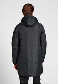 Man som står med ryggen mot kameran iförd en lång svart kapuschongjacka med vadderade ärmar och svarta byxor mot en neutral vit bakgrund.