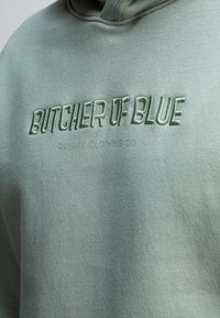 Groene hoodie met geborduurde tekst "BUTCHER OF BLUE" en "QUALITY CLOTHING CO" in bijpassend garen. Zachte, gestructureerde stof met een relaxte pasvorm.