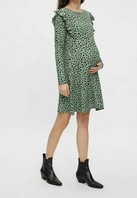 Zelené šaty s dlouhými rukávy, černými puntíky, volány na ramenou a rozšířeným lemem, doplněné černými kotníkovými botami.