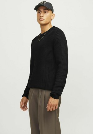 Maglione nero lavorato a maglia con scollatura rotonda e texture a coste, abbinato a pantaloni beige plissettati. Indossato con un cappellino scuro e una collana dorata.
