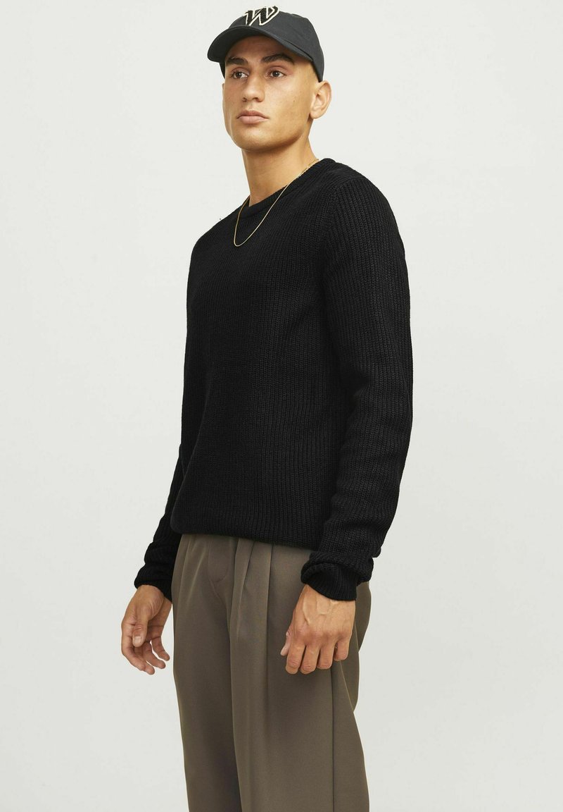 Maglione nero lavorato a maglia con scollatura rotonda e texture a coste, abbinato a pantaloni beige plissettati. Indossato con un cappellino scuro e una collana dorata.