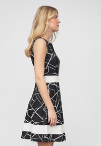 Robe noire avec un motif géométrique blanc, sans manches, corsage ajusté, jupe évasée et ceinture blanche contrastante.