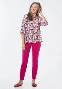 Bunte gemusterte Bluse mit geometrischen Formen, kombiniert mit hellrosa, enganliegenden Hosen und rosa Schlüpfschuhen. Glattes Material, lässige Passform.