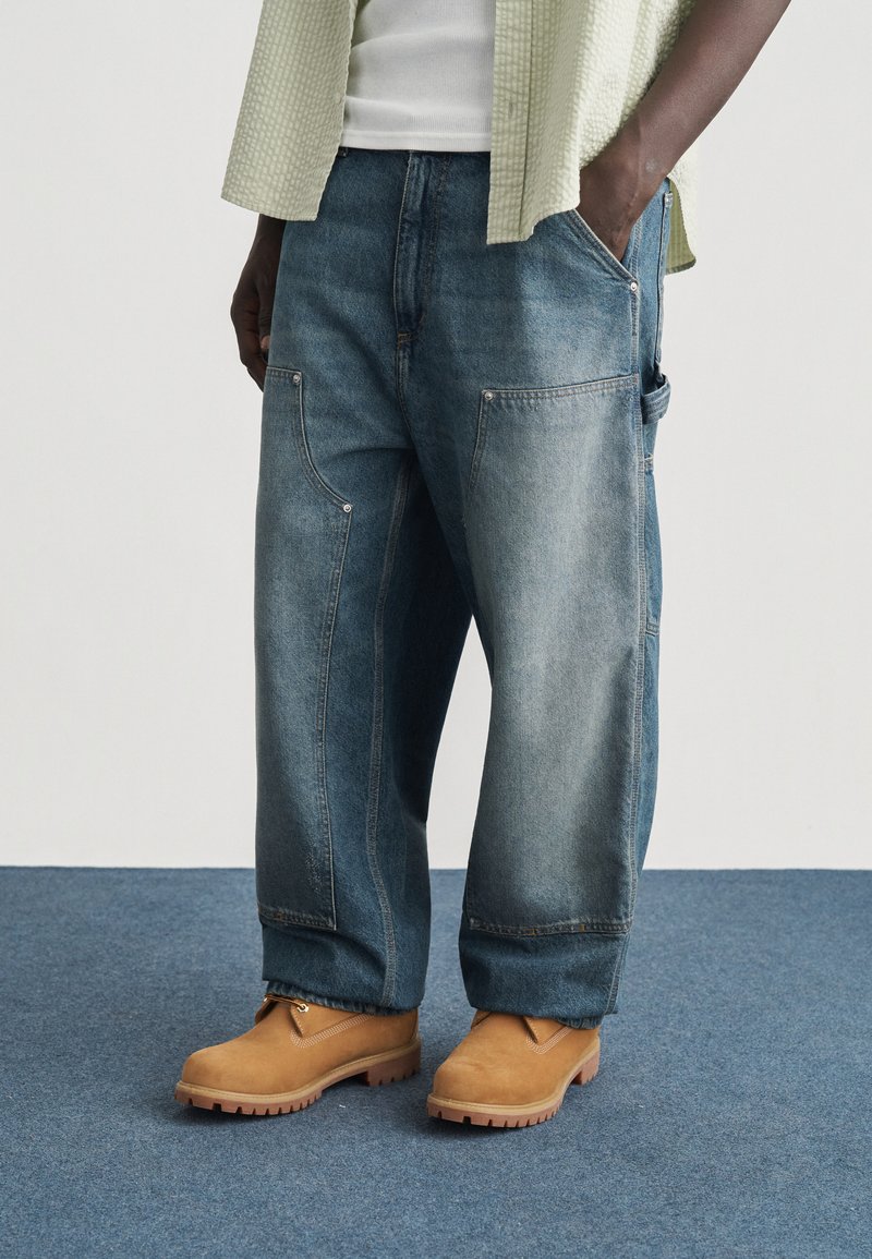 Άτομο που φοράει φαρδιά μπλε τζιν παντελόνια με μεγάλες τσέπες, ταμπά εργαστικές μπότες, λευκό πουκάμισο και ανοιχτό πράσινο υφασμάτινο επάνω πουκάμισο.